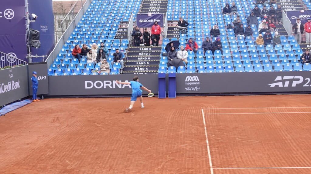 Young Romanian tennis player Stefan Horia Haita hits a backhand while spectators with umbrellas watch from the stands at the Tiriac Open
