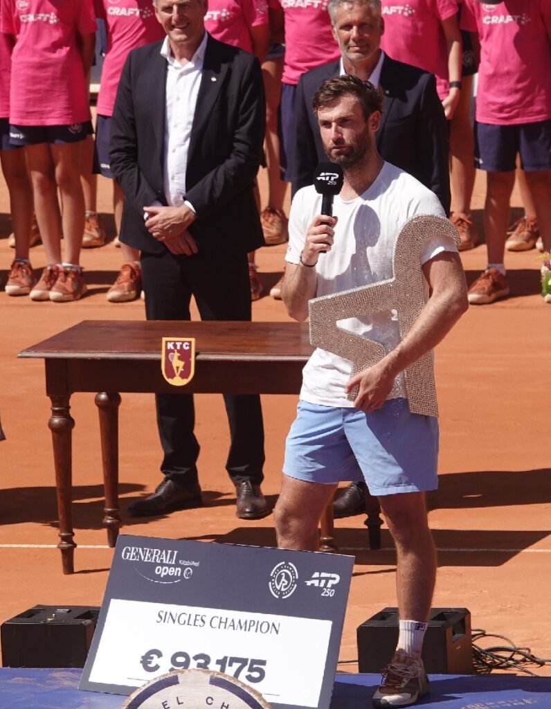 Quentin Halys giving his victory speech while holding the trophy and standing behind the prize money check at the 2026 ATP Kitzbuhel tournament.