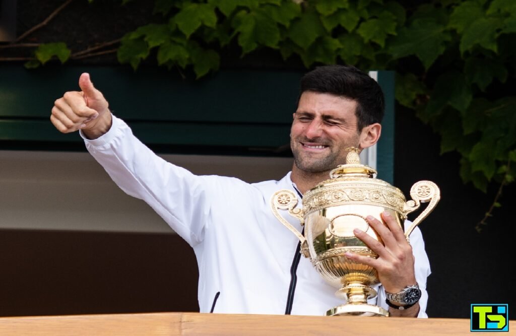 Novak Djokovic smiling and giving a thumbs up while holding the Wimbledon trophy, celebrating a major victory at the tournament.