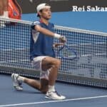 Le joueur de tennis japonais Rei Sakamoto à genoux sur le court près du filet célébrant sa victoire en tenant sa raquette comme une épée de samouraï à l'ATP Almaty Open.
